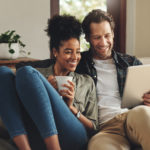 A young couple sitting on the sofa, looking at a tablet and smiling.