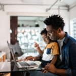 A father holding a son on his lap and using a computer.